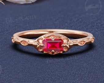 Op de afbeelding: Een roségouden ring met een rechthoekige, rode edelsteen in het midden. De ring heeft een delicaat bloemmotief op de band en rond de edelsteen. De ring is geplaatst tegen een donkerblauwe achtergrond.