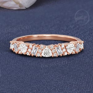 May include: Rose gold ring with a row of diamonds. The ring features a mix of round, baguette, and pear-shaped diamonds. The ring is set against a dark blue background, with a white rose in the background. The text "BlessGem" is visible.