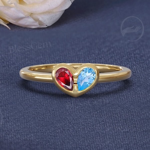 May include: A gold ring with a heart-shaped design featuring a red and blue gemstone. The ring is on a blue fabric background.