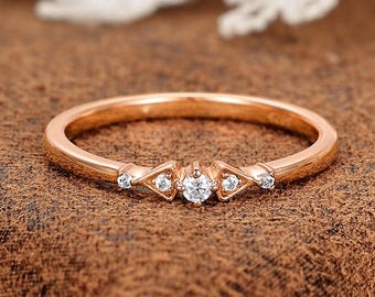 May include: A rose gold ring with a delicate band. The ring features a central round diamond flanked by two diamond-shaped settings, each with a smaller diamond. The ring is set against a brown textured surface.