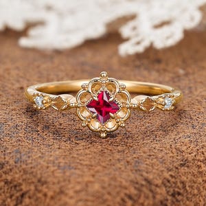 May include: A gold ring with a square, deep red gemstone at the center, surrounded by a decorative floral design. The band features small diamond accents and intricate detailing. The ring is set against a brown background.