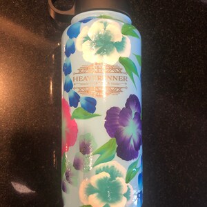 May include: A light blue water bottle with a floral design featuring white, blue, and purple flowers. The bottle has a black lid and a black handle. The text "HEAVY RUNNER" is printed in gold on the bottle.