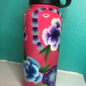 May include: Pink stainless steel water bottle with a black lid and handle. The bottle is decorated with hand-painted purple, blue and white flowers.