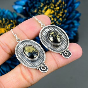 May include: A pair of silver earrings featuring oval black and gold gemstones. The earrings have a textured, dotted border and a decorative swirl. The earrings are displayed against a dark background.