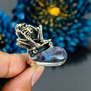 Op de afbeelding: Een zilveren ring met een blauwe en roze edelsteen en een gouden skeletfiguur dat op een skateboard zit.