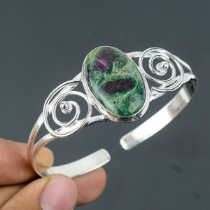 May include: A silver bangle bracelet featuring a green and pink oval gemstone set in the centre. The bracelet has a delicate, swirling design.
