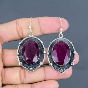 May include: A pair of silver earrings with a large oval-shaped purple gemstone set in a silver frame. The frame has a textured finish and a small flower design at the bottom.
