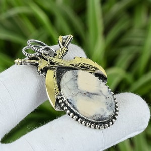 Puede incluir: Un colgante de plata y latón con una piedra blanca y gris rodeada de dos espadas cruzadas. Las espadas tienen detalles intrincados y están colocadas sobre un fondo de latón.