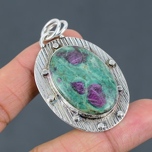 Könnte beinhalten: Ovaler Anhänger mit einem grün-rosa Edelstein, der in einen silbernen Rahmen gefasst ist. Der Rahmen hat ein strukturiertes Design und ist mit kleinen silbernen Punkten verziert.