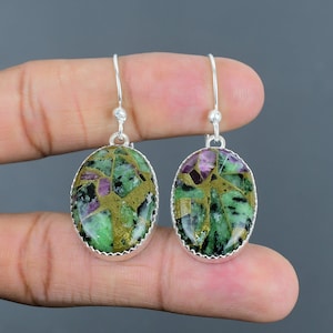 May include: A pair of oval earrings with a silver setting. The earrings display a mosaic of green, gold, and purple stones. The earrings are suspended by a silver hook.