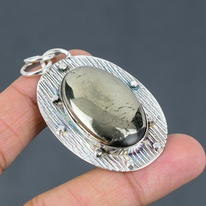 Puede incluir: Pendiente de plata con una gran piedra ovalada de pirita engastada en un marco de plata texturizado. El pendiente tiene una anilla de plata para sujetarlo a una cadena.