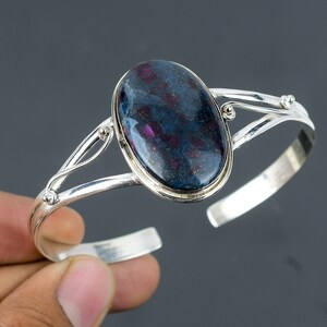 May include: A silver bangle bracelet with a large oval-shaped blue gemstone with pink flecks. The bracelet has a delicate, twisted design.
