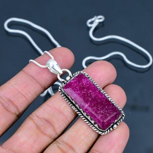 May include: A rectangular pendant featuring a vibrant magenta gemstone set in a silver frame with a twisted rope border. The pendant hangs from a thin silver snake chain, showcasing a polished metallic finish and a textured, faceted stone surface.