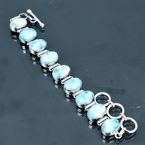 Puede incluir: Una pulsera de plata con piedras preciosas ovaladas de color azul claro. La pulsera tiene un cierre de palanca y está compuesta por múltiples eslabones de piedras preciosas. Las piedras tienen un aspecto veteado.