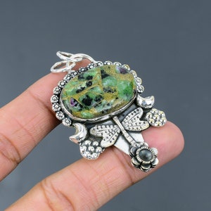 Puede incluir: Un colgante de plata con una piedra preciosa verde y dorada engastada en un diseño de libélula de plata. El colgante está en un dedo.
