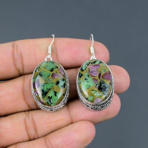 May include: A pair of oval earrings with a silver metal setting. The earrings display a mosaic of green, gold, and purple stones. The earrings are suspended from a silver hook.