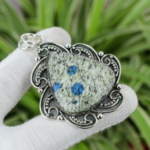 Könnte beinhalten: Ein silberner Anhänger mit einem blau-weiß gefleckten Stein, der in einen dekorativen silbernen Rahmen gefasst ist. Der Anhänger hat eine Öse zum Anbringen einer Kette.