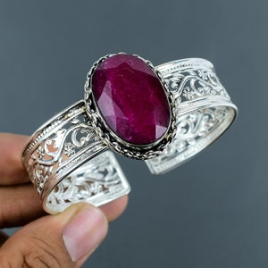 May include: A silver bangle bracelet with an ornate design features a large, oval-shaped, faceted ruby gemstone.