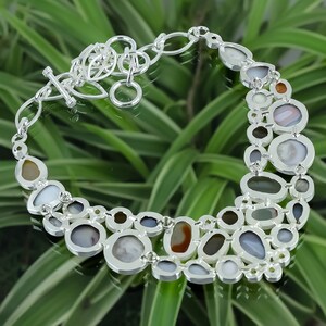 May include: A silver necklace with a unique design featuring a variety of oval-shaped stones in shades of brown, gray, and white. The stones are set in silver and are connected by a silver chain.