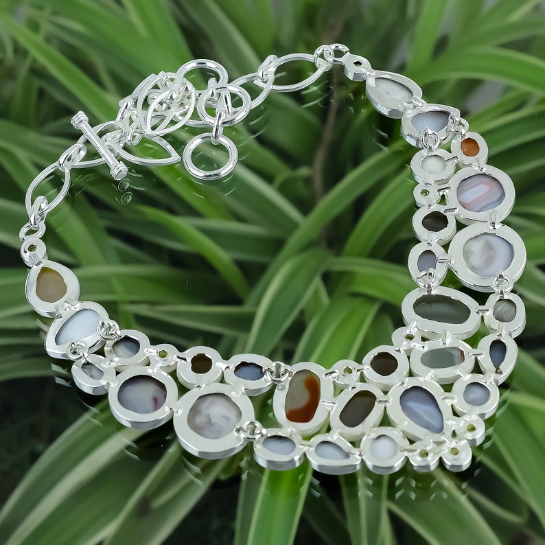 May include: A silver necklace with a unique design featuring a variety of oval-shaped stones in shades of brown, gray, and white. The stones are set in silver and are connected by a silver chain.