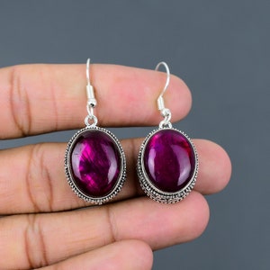 Puede incluir: Un par de pendientes de plata con piedras ovaladas de color morado oscuro. Los pendientes tienen un diseño de plata detallado alrededor de los bordes de las piedras preciosas.