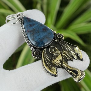 May include: A silver pendant with a blue gemstone and a gold dragon design. The pendant is on a white surface.