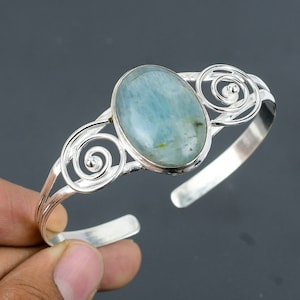 Puede incluir: Un brazalete de plata con una piedra ovalada azul claro engastada en el centro. El brazalete tiene un diseño delicado y en espiral.