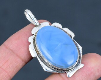 May include: A light blue oval pendant with a silver-colored bail and decorative frame. The pendant is held by a hand against a dark background. The gemstone has a smooth, polished surface.