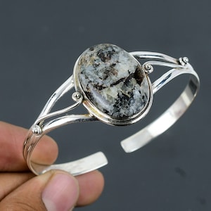 Op de afbeelding: Een zilveren armband met een grote ovale zwarte en witte edelsteen. De armband heeft een delicaat, opengewerkt ontwerp.