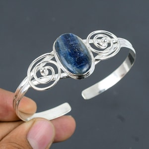 May include: A silver cuff bracelet featuring a central oval-shaped blue kyanite gemstone. The metal band is accented with symmetrical wire-work spiral designs on both sides of the stone, creating a polished and reflective metallic finish.