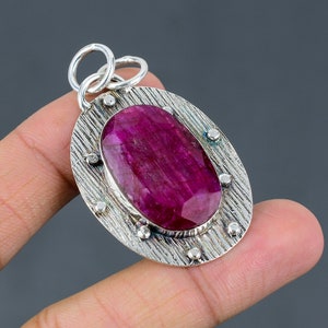 May include: A silver pendant featuring a large oval ruby gemstone set within a textured silver frame. The pendant has a loop at the top for attaching a chain.