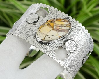 May include: A silver cuff bracelet with a large oval-shaped gemstone set in the center. The gemstone is a mix of white, yellow, and brown colors. The bracelet has a textured surface and is decorated with cut-out flower shapes.