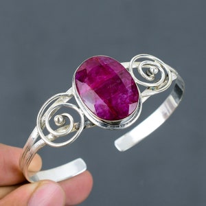 Pode incluir: Um bracelete de prata com uma grande gema de rubi ovalada engastada no centro. O bracelete apresenta três volutas decorativas de cada lado da gema.
