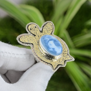 Puede incluir: Anillo de plata con una piedra preciosa ovalada de color azul claro. El anillo tiene un engaste decorativo de color dorado y un diseño detallado con patrones alrededor de la piedra preciosa, que se asemeja a una forma de tortuga.