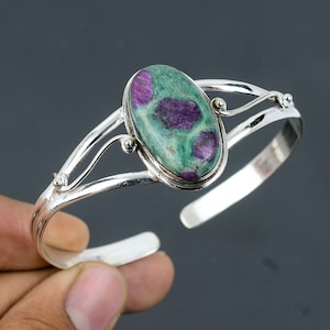 Könnte beinhalten: Ein silbernes Armreif-Armband mit einem ovalen grünen und violetten Edelstein, der in der Mitte eingesetzt ist. Das Armband hat ein zartes, geschwungenes Design.