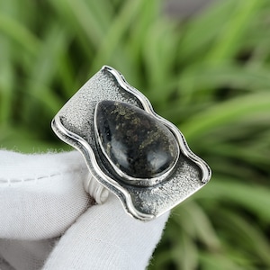 Könnte beinhalten: Ein silberner Ring mit einem dunkelgrünen Edelstein, der in einem strukturierten, wellenförmigen Design gefasst ist.