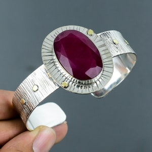 Könnte beinhalten: Ein silbernes Armreif-Armband mit einem großen, ovalen Rubin-Edelstein, der in einer strukturierten Silberfassung sitzt. Das Armband hat eine gehämmerte Oberfläche und goldene Akzente.