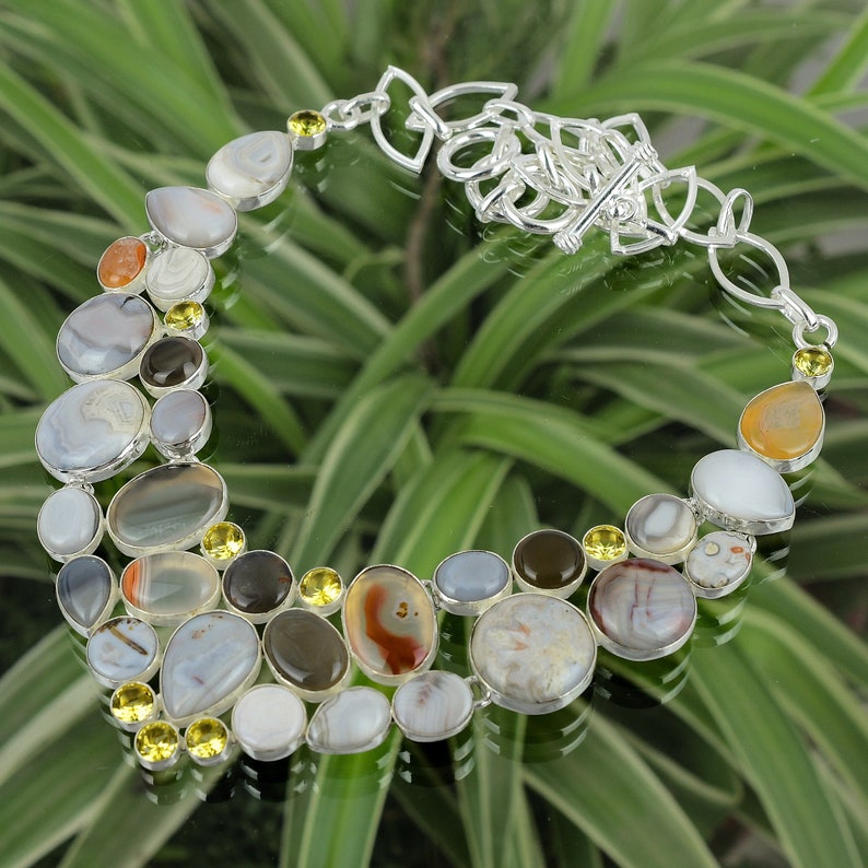 May include: A silver necklace with a clasp closure features a variety of agate stones in shades of gray, brown, and white. The stones are set in silver and are interspersed with small yellow stones.