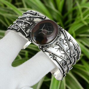 May include: A silver cuff bracelet with a large, oval-shaped, brown and black gemstone set in the center. The bracelet is decorated with a repeating pattern of small silver dots and lines.