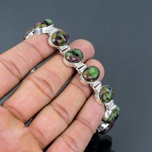 May include: A silver bracelet featuring oval-shaped gemstones with a mix of green, purple, and gold hues. The gemstones are set in individual silver bezels and linked together with silver bars. The bracelet is displayed against a dark background.