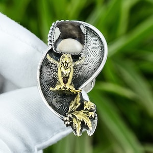 May include: A silver ring with a large, rectangular, black and white stone. The ring features a gold-colored owl and leaf design. The ring has intricate detailing and is set against a green background.