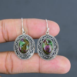 May include: Silver earrings with oval gemstones in shades of green, purple, and gold. The earrings have a decorative, filigree-style border. The gemstones are the focal point of the design.