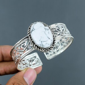 Puede incluir: Un brazalete de plata con una piedra ovalada de howlita blanca. El brazalete tiene un delicado diseño de filigrana.