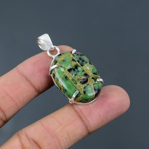 Puede incluir: Un colgante de forma ovalada que presenta una piedra verde y negra con vetas de color dorado metálico, engastado en una montura de metal plateado pulido con garras y una anilla plateada a juego en la parte superior para una cadena.