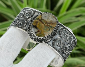 May include: A wide silver cuff bracelet with an oval, multi-colored stone at the center. The stone features shades of brown, green, and cream. The bracelet has an ornate, swirling design and is approximately 2 inches wide.