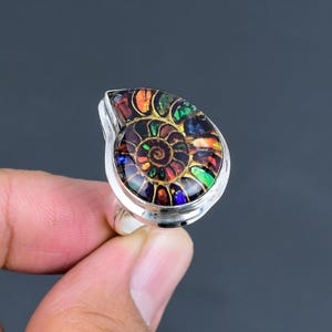 May include: A silver ring with an iridescent ammonite fossil gemstone. The fossil has a spiral design with red, green, yellow, and blue colours. The ring has a polished silver band and setting, highlighting the gemstone.