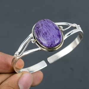 Pode incluir: Um bracelete de prata com uma grande pedra preciosa ovalada de cor púrpura engastada em um bezel de prata. O bracelete tem um design delicado e vazado.