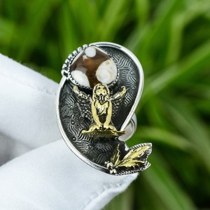 May include: A silver ring with a brown and white stone set in a heart-shaped design. The ring features a gold angel figure with outstretched wings.
