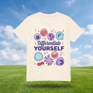 Op de afbeelding: Crèmekleurig T-shirt met kleurrijke illustraties van cellen en de tekst "Differentiate YOURSELF". Het ontwerp bevat verschillende celtypen in tinten roze, blauw, paars en rood, met ster- en diamantaccenten. Het shirt wordt gepresenteerd tegen een achtergrond van een blauwe lucht en groen gras.