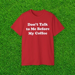 May include: A bright red short-sleeved t-shirt featuring the white text "Don't Talk to Me Before My Coffee" printed in a bold, sans-serif font across the center of the chest. The garment has a classic crew neckline and a simple, straight hem.